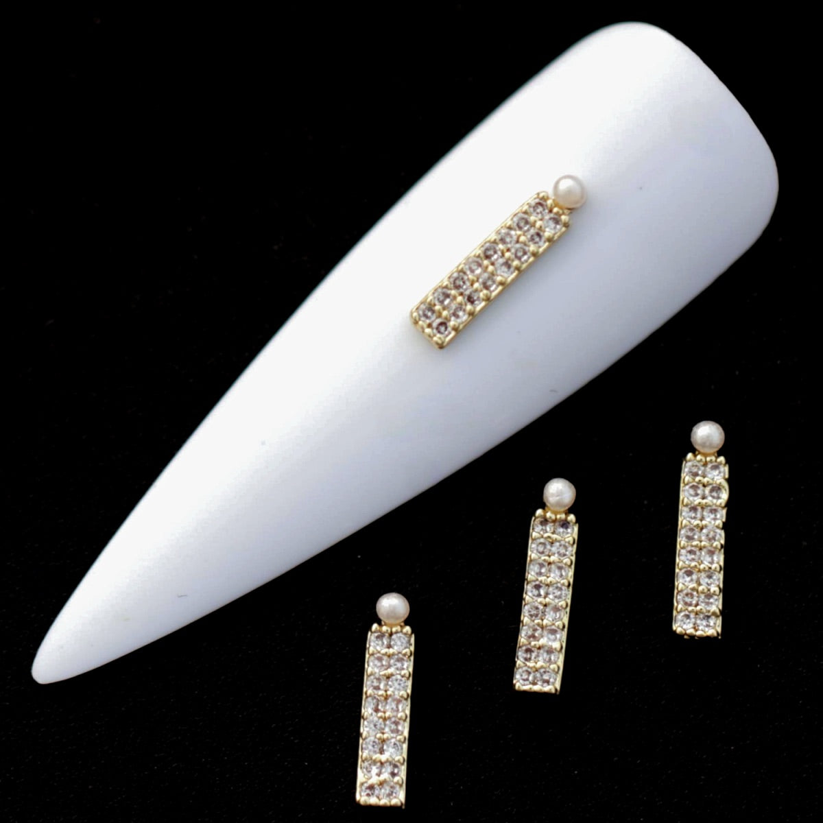 Gold bar nail charms paved with crystal CZ stones and topped with small half‑pearls on a white nail tip against a black background.