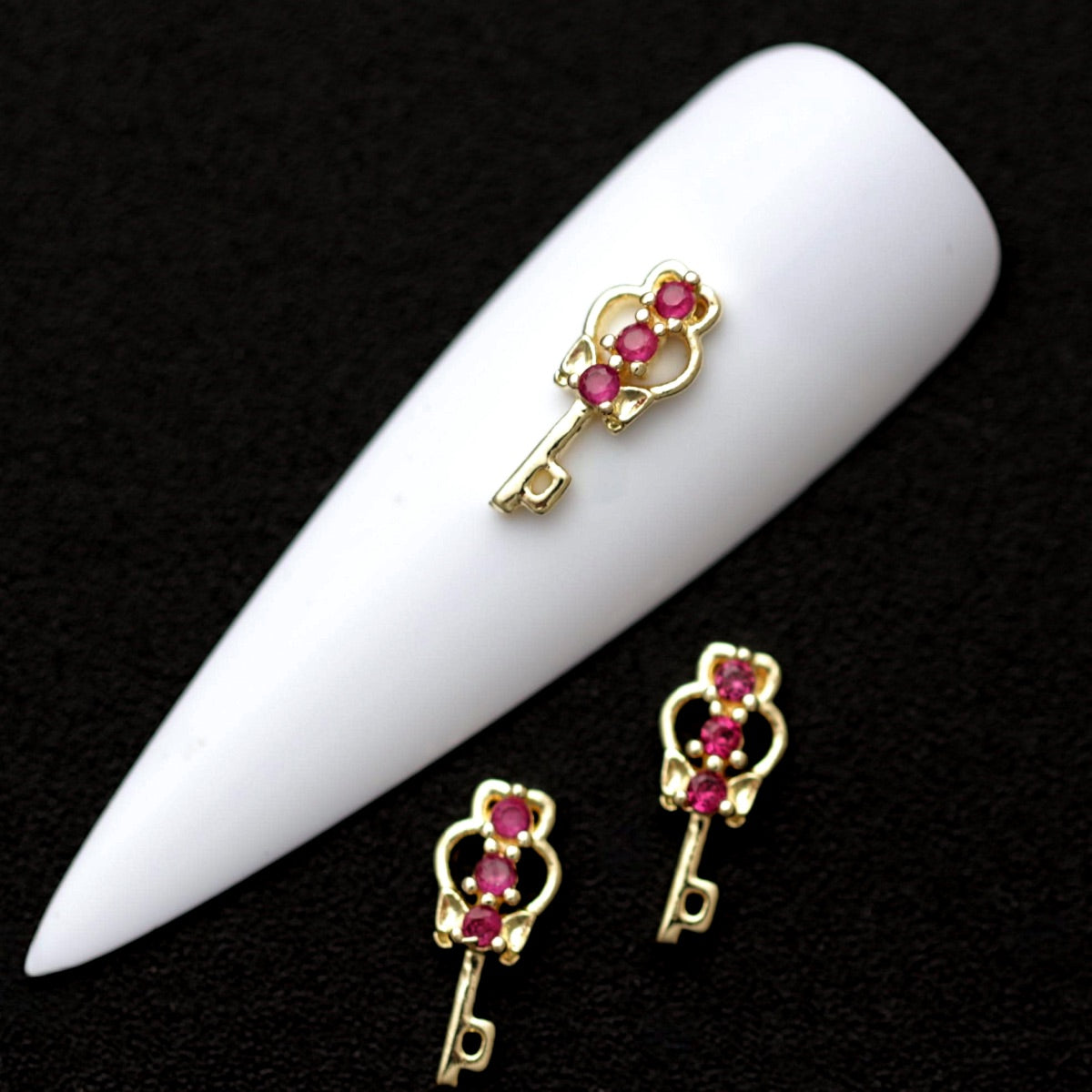 Gold 11×7 mm key-shaped nail charms with magenta CZ stones displayed on a white nail tip against a black background