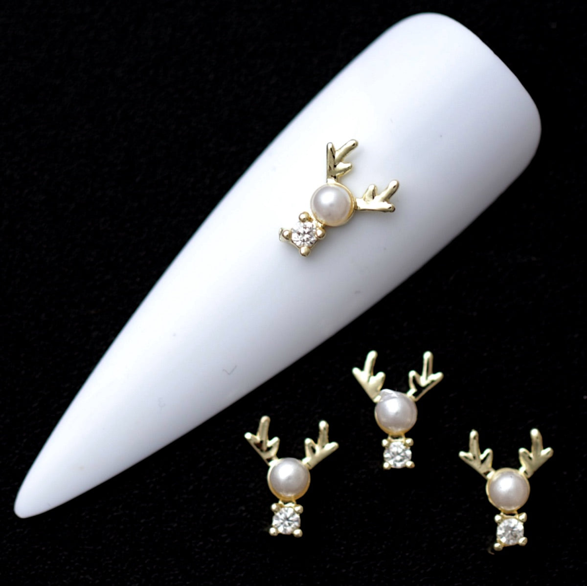 Gold reindeer nail charms with white pearl heads and crystal CZ noses on a white nail tip against a black background