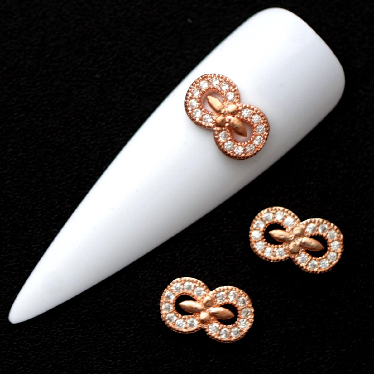 Rose gold infinity nail charms paved with crystal CZ stones on a white nail tip against a black background