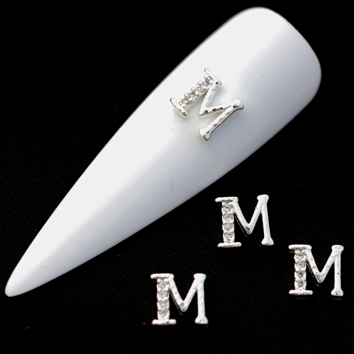 Silver M letter nail charms with crystal CZ accents on a white nail tip against a black background.