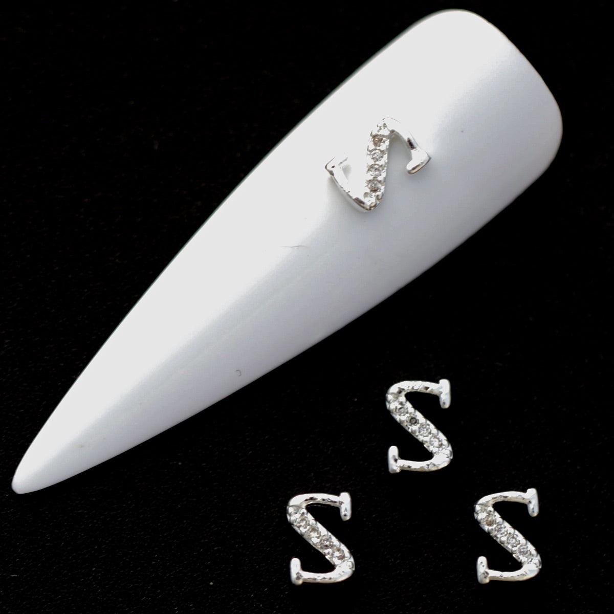 Silver S letter nail charms with crystal CZ accents on a white nail tip against a black background.
