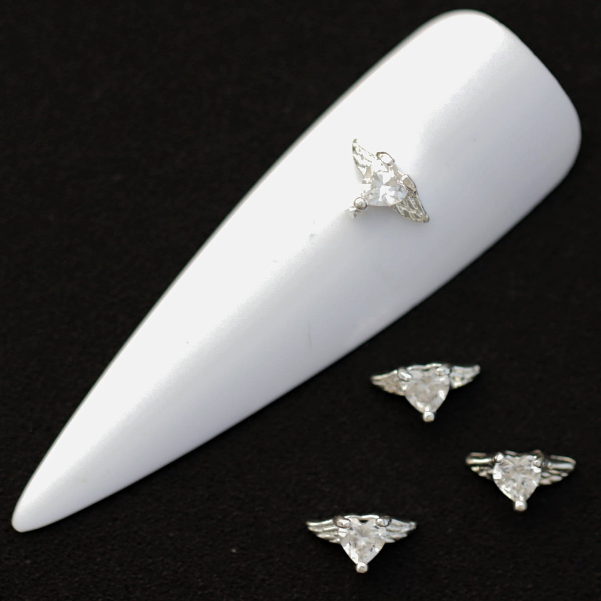 Silver winged heart nail charms with crystal‑colored heart CZ stones on a white nail tip against a black background