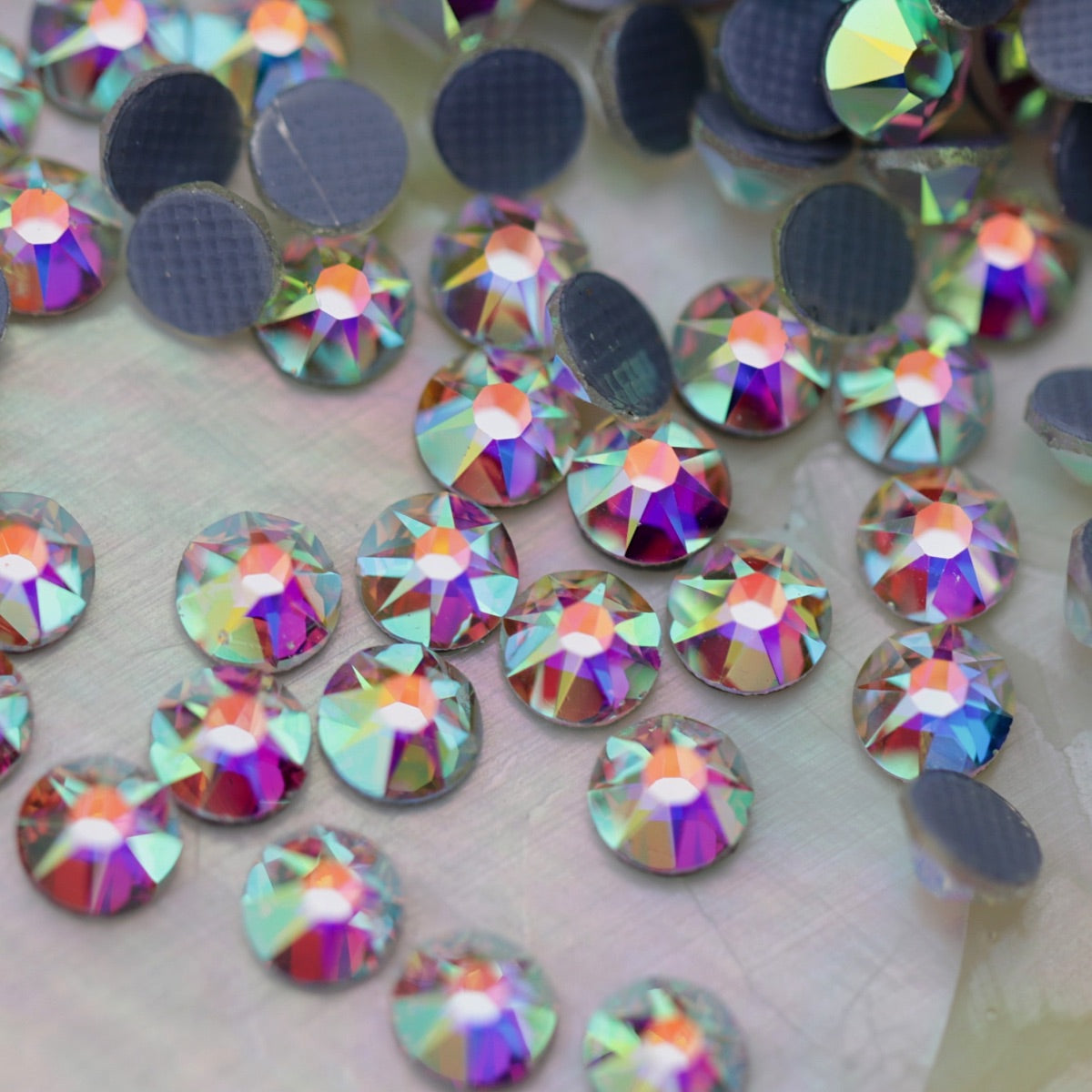 Close-up of round Crystal AB Eunails hotfix rhinestones with faceted tops and grey textured hotfix backs scattered on a light surface.
