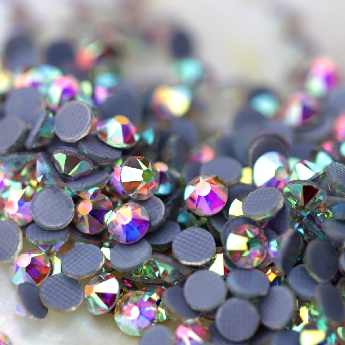 Close-up of round Crystal AB Eunails hotfix rhinestones with faceted tops and grey textured hotfix backs scattered on a light surface.