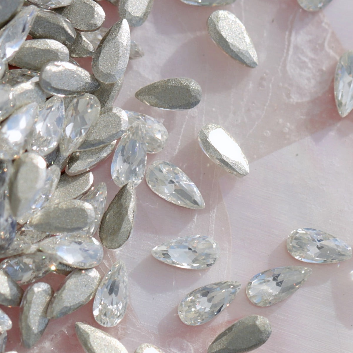 Pile of metallic silver drop-shaped flatback zircon crystals on pearlescent pink background, perfect for elegant nail art embellishments