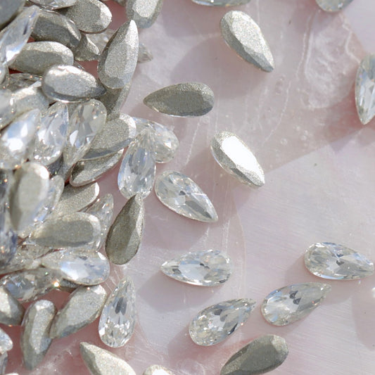 Pile of metallic silver drop-shaped flatback zircon crystals on pearlescent pink background, perfect for elegant nail art embellishments
