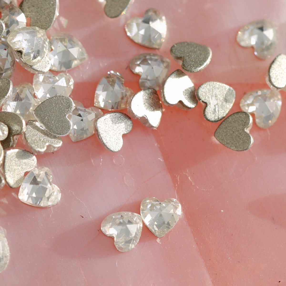 Scattered heart-shaped cubic zirconia flatback crystals showing faceted sparkle and metallic silver base on soft pink surface for nail decoration