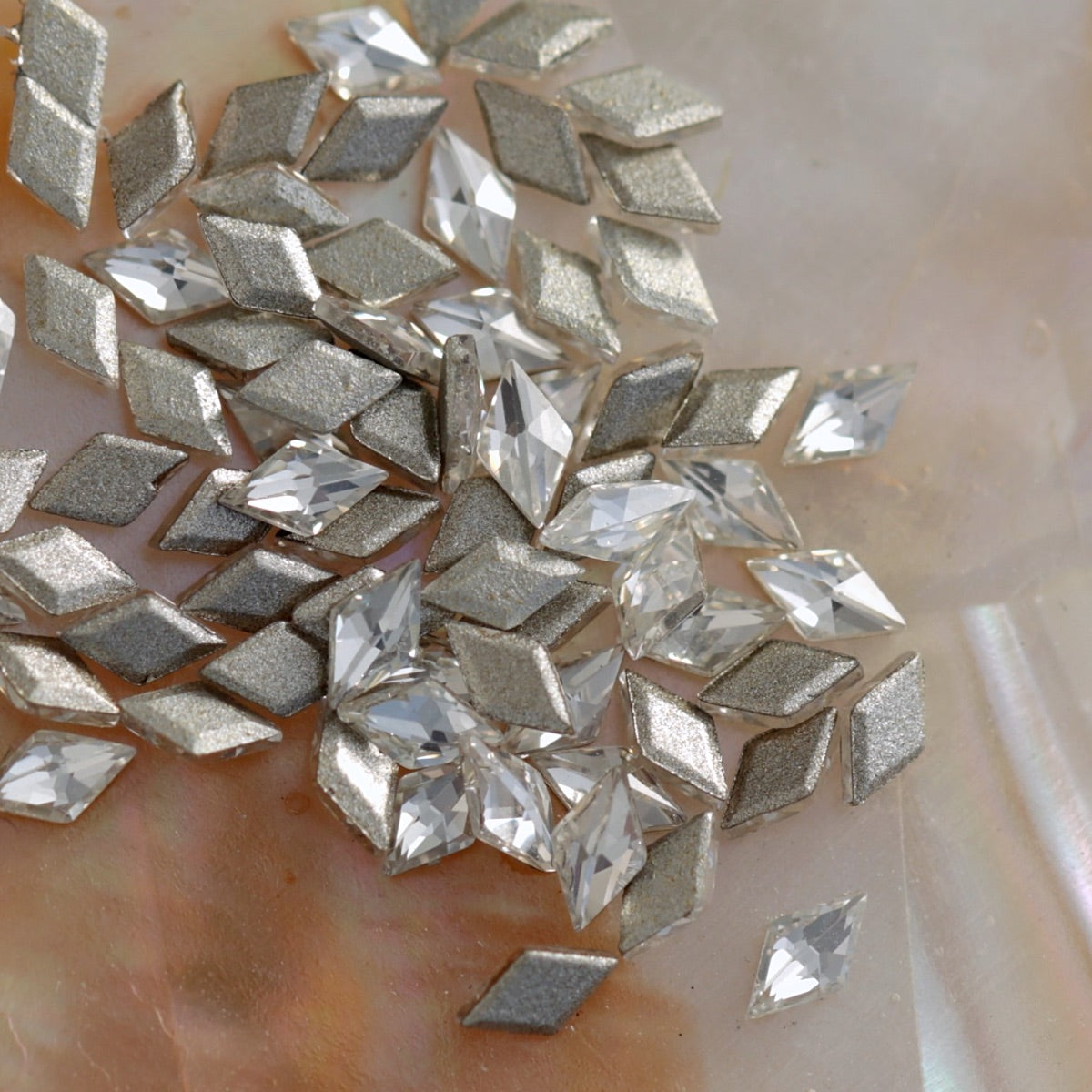 Flatback zircon rhombus crystals in silver and clear tones scattered on pearl background, perfect for nail art decoration