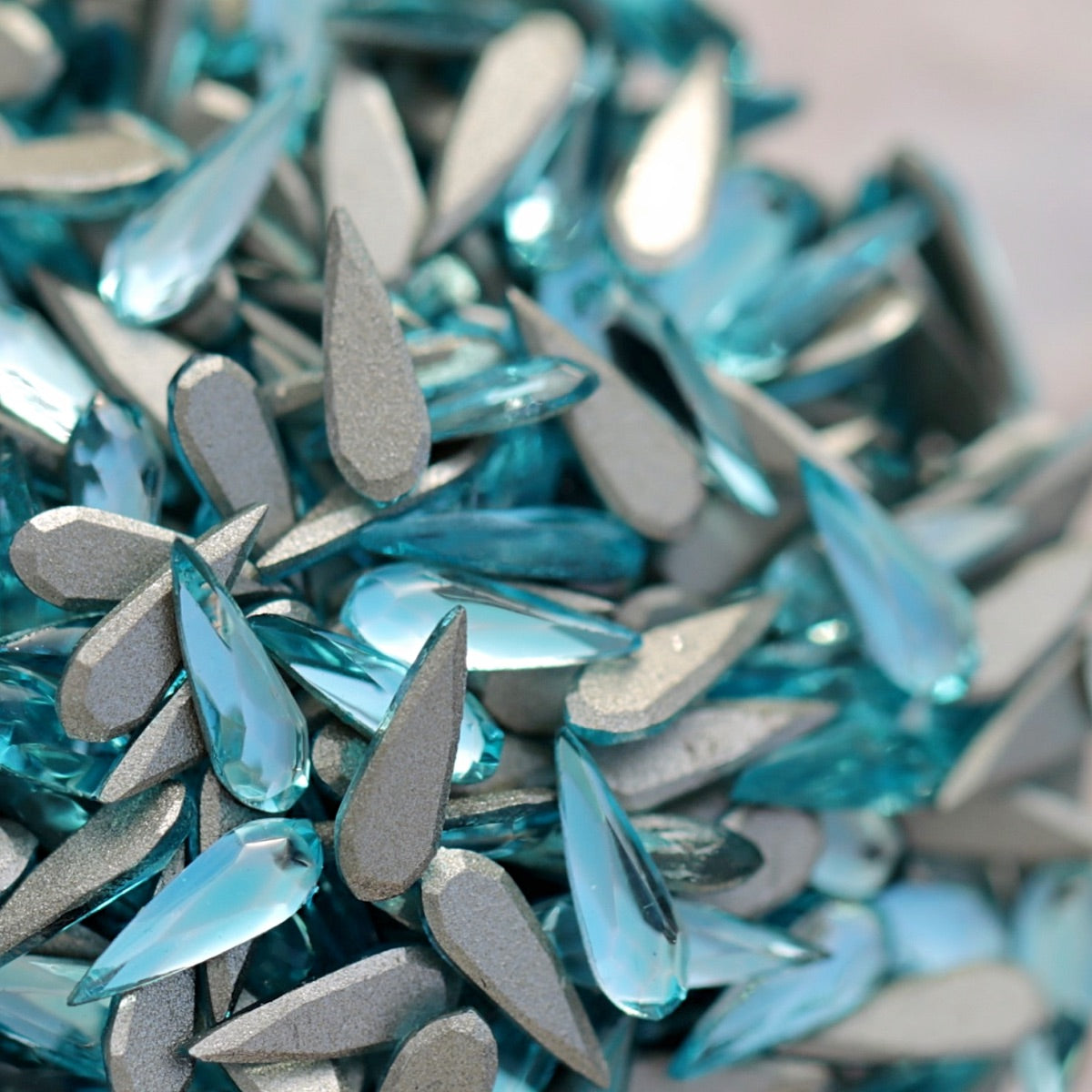 Pile of aquamarine glass flatback raindrop rhinestones with silver foil backing and faceted sparkle on light background for nail art