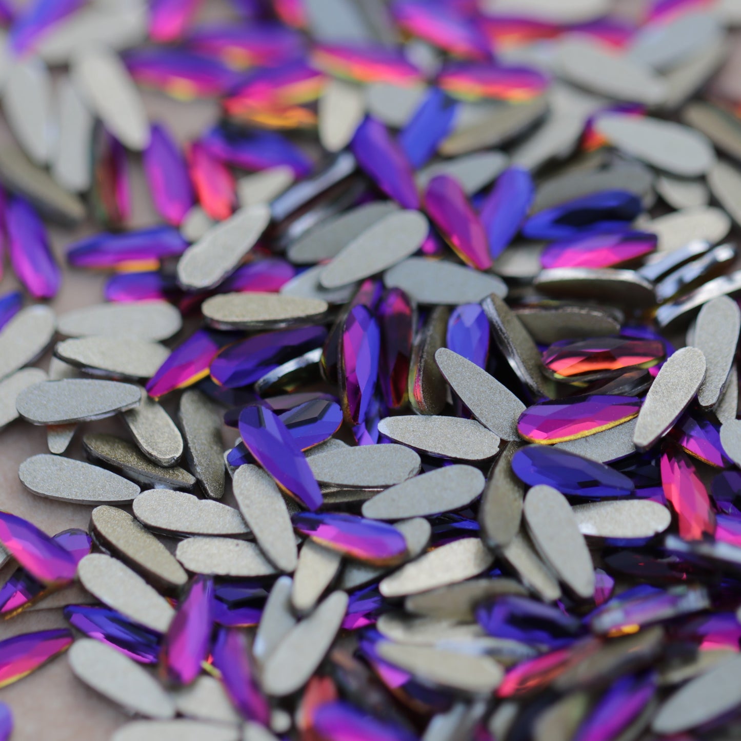 Slim flatback raindrop glass rhinestones in Meridian Blue showing blue, violet and pink reflections scattered on a surface.