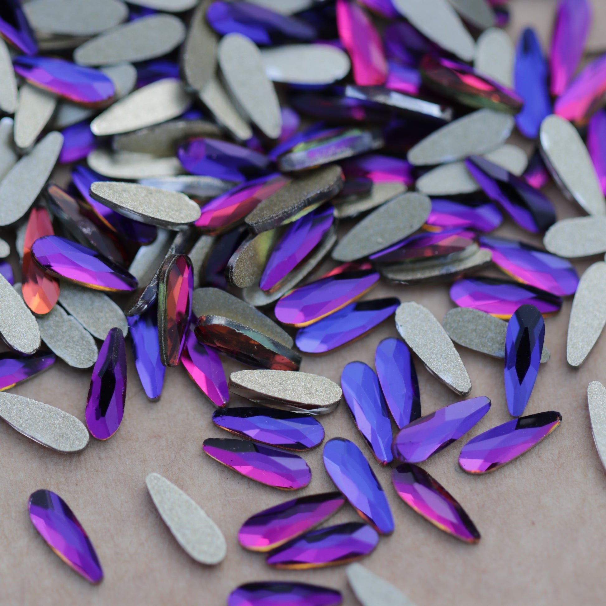 Slim flatback raindrop glass rhinestones in Meridian Blue showing blue, violet and pink reflections scattered on a surface.