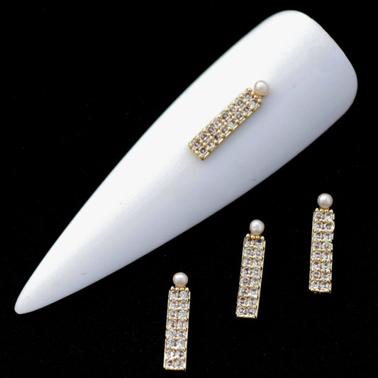 Gold bar nail charms paved with crystal CZ stones and topped with small half‑pearls on a white nail tip against a black background.