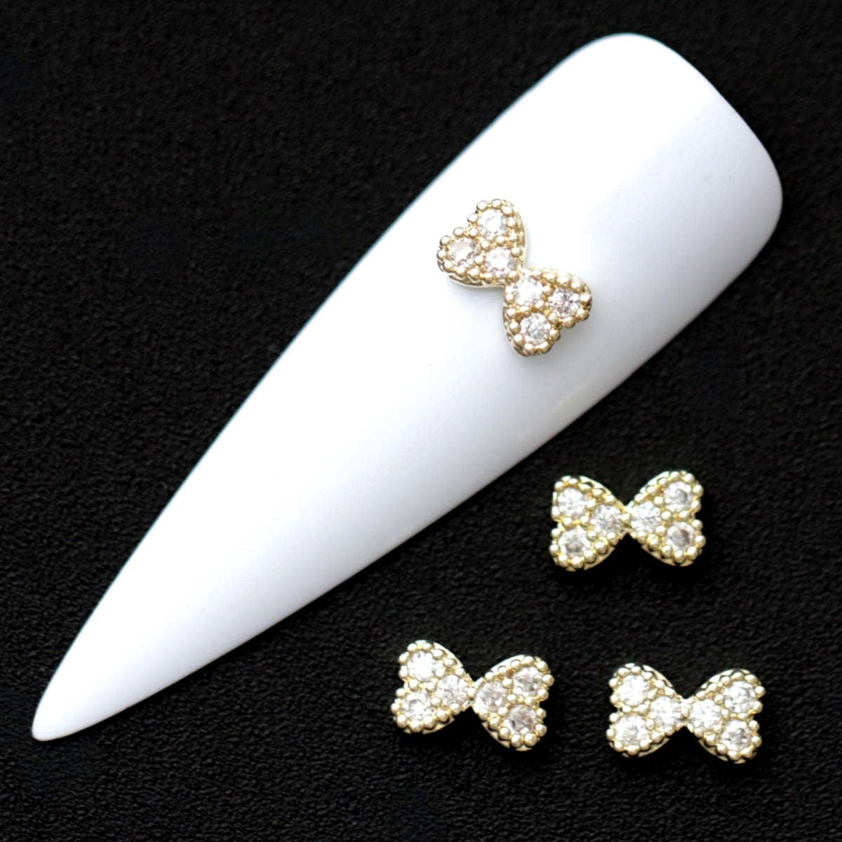 Gold 7×5 mm bow‑shaped nail charm fully paved with mini crystal CZ stones displayed on a white nail tip against a black background