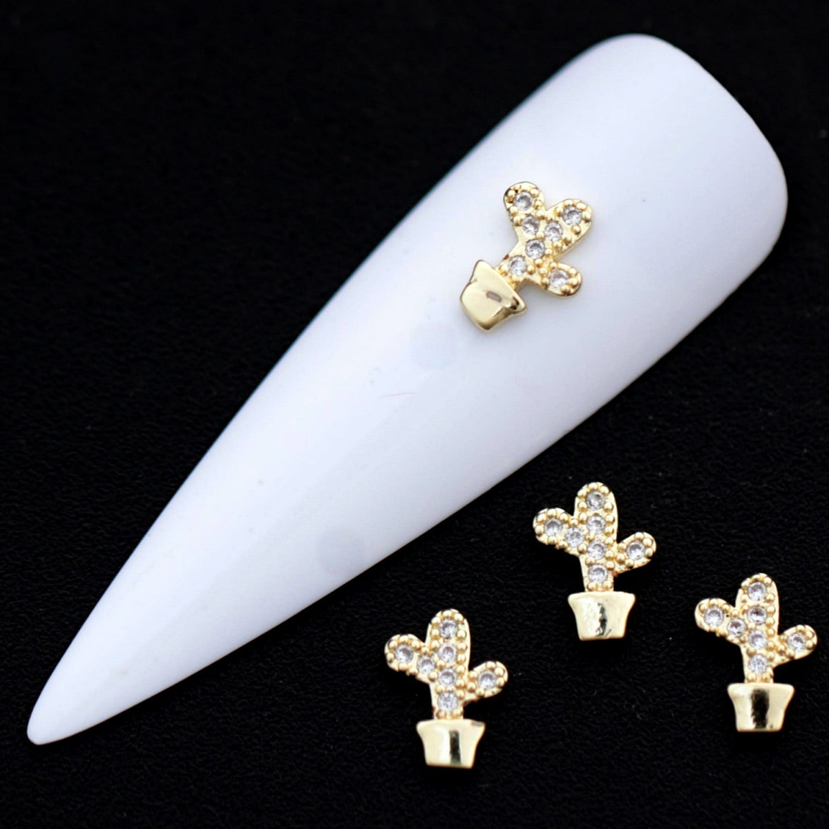 Gold potted cactus nail charms with crystal CZ stones on a white nail tip against a black background.