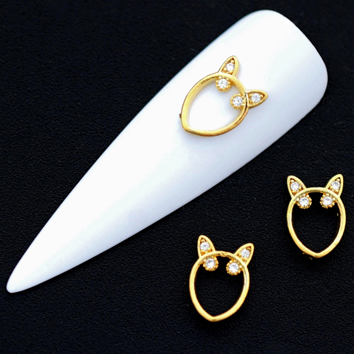 Gold cat‑head frame nail charms with crystal‑colored CZ accents on a white nail tip against a black background.