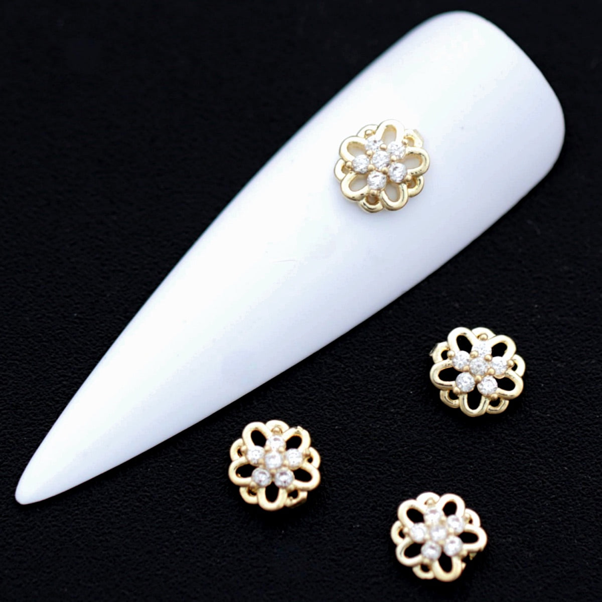 Gold flower‑shaped nail charms with crystal‑colored CZ stones on a white nail tip against a black background.