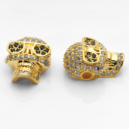 Large gold skull beads paved with crystal and jet CZ stones shown on a white nail tip against a white background.