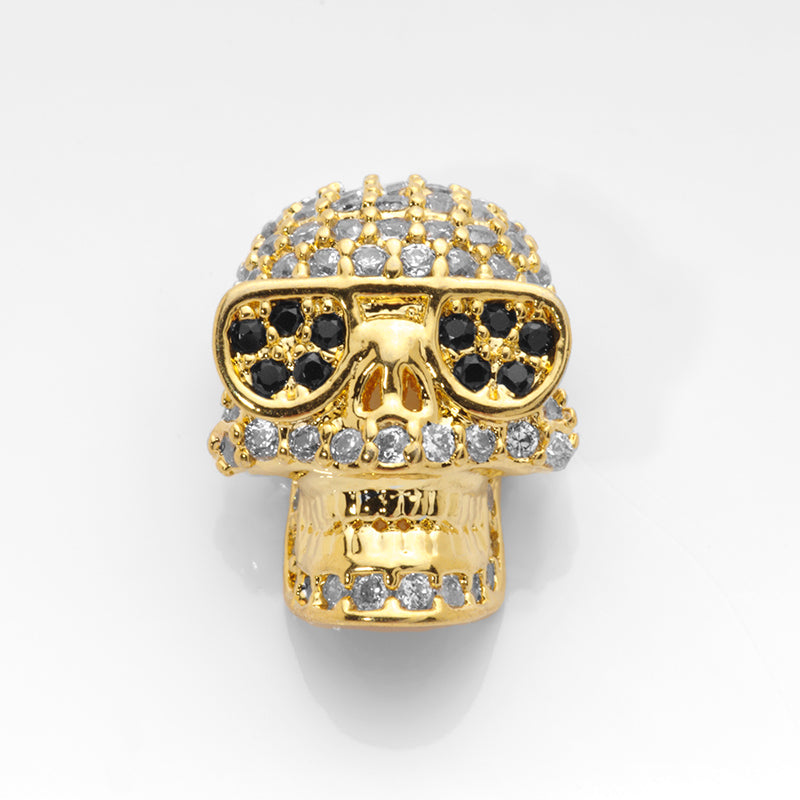 Large gold skull beads paved with crystal and jet CZ stones shown on a white nail tip against a white background.