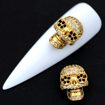 Large gold skull beads paved with crystal and jet CZ stones shown on a white nail tip against a black background.