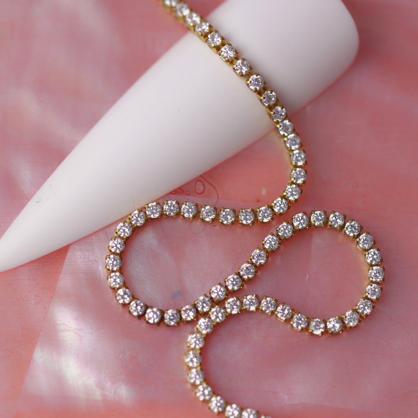 Gold 1.2 mm clear zirconia rhinestone nail art chain wrapped around a white nail tip on a pink background