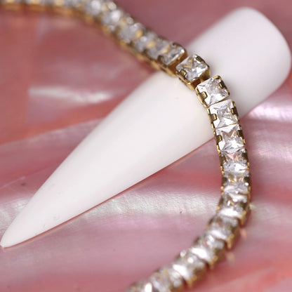 Gold cup chain with 3×3 mm clear zirconia square rhinestones wrapped around a white nail tip on a pink background