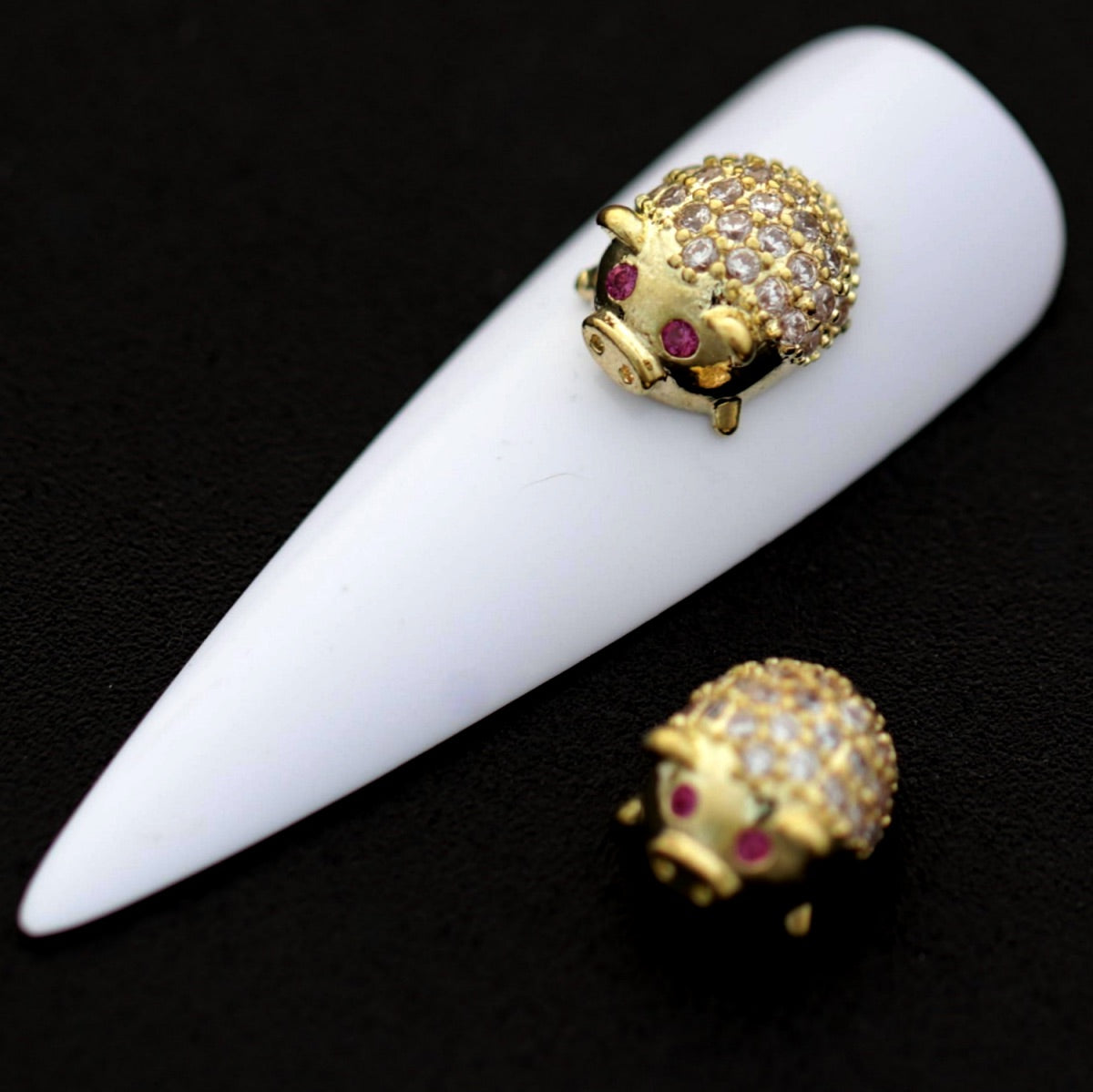Gold lucky pig nail charms with crystal CZ backs and pink CZ eyes shown on a white nail tip against a black background.