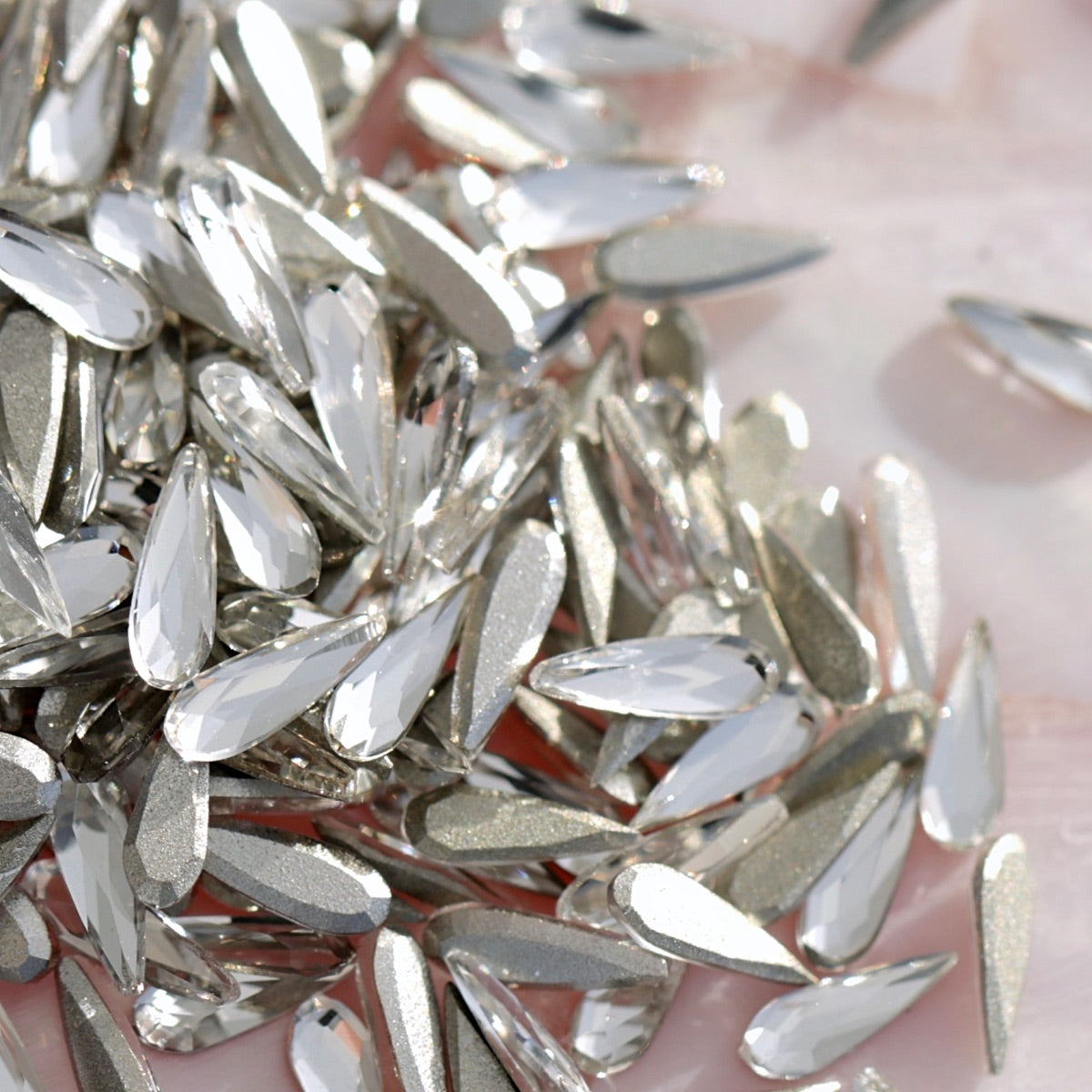 Pile of clear K9 crystal flatback raindrop shaped rhinestones with metallic silver tones on pearlescent pink background for nail art