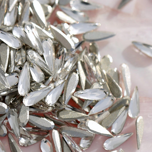 Pile of clear K9 crystal flatback raindrop shaped rhinestones with metallic silver tones on pearlescent pink background for nail art