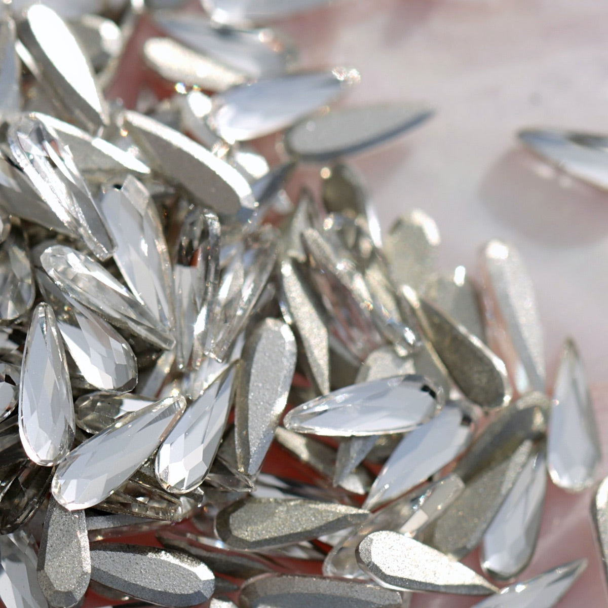 Scattered clear raindrop K9 flatback crystals showing faceted sparkle and silver metallic backing on soft surface for nail decoration