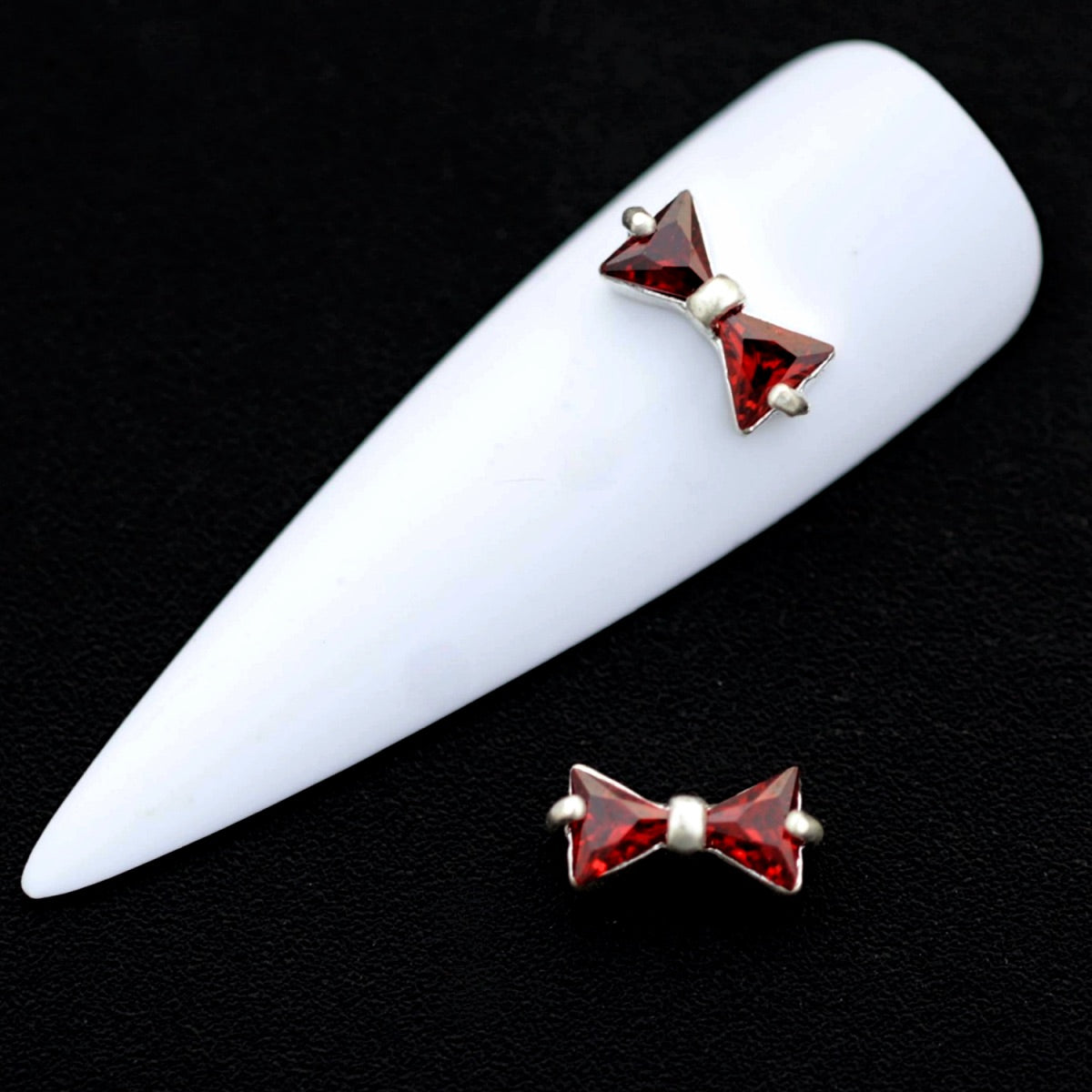 Silver bow‑shaped nail charms with amber red CZ stones on a white nail tip against a black background.