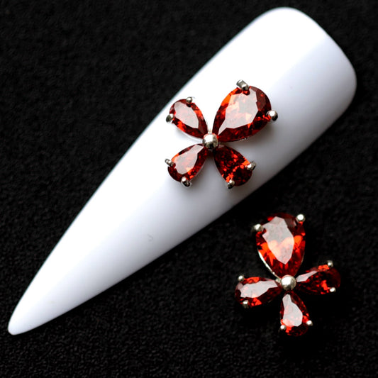 Silver ankh‑style nail charms with large amber‑red pear‑cut CZ stones arranged in a cross shape on a white nail tip against a black background