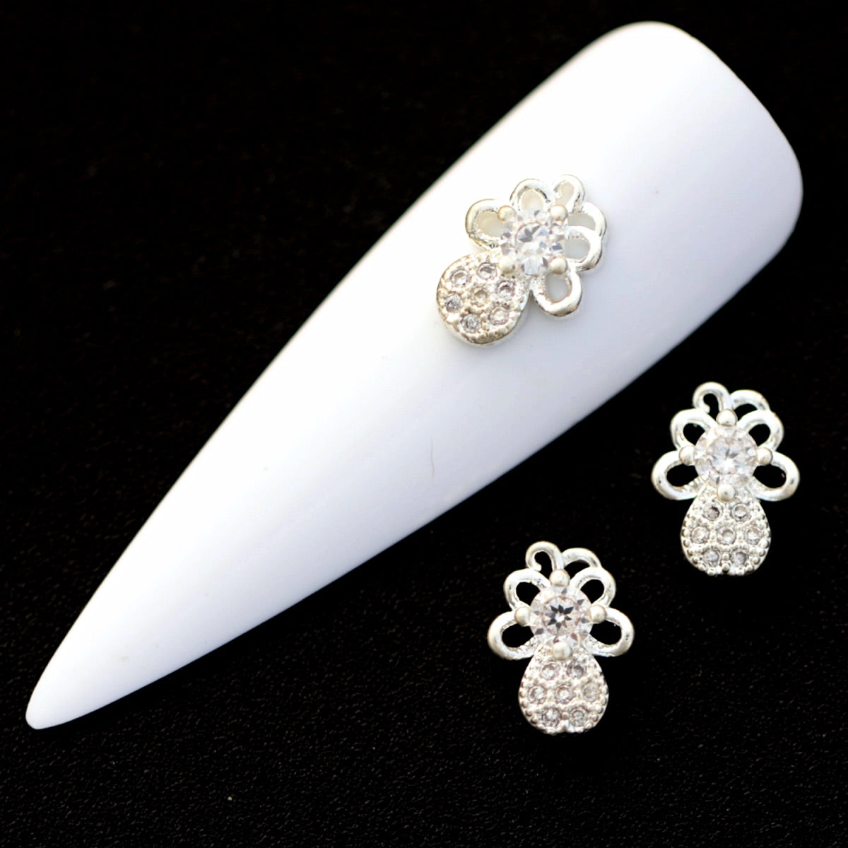 Silver bow nail charms with central round crystal CZ and pavé teardrop tails on a white nail tip against a black background.