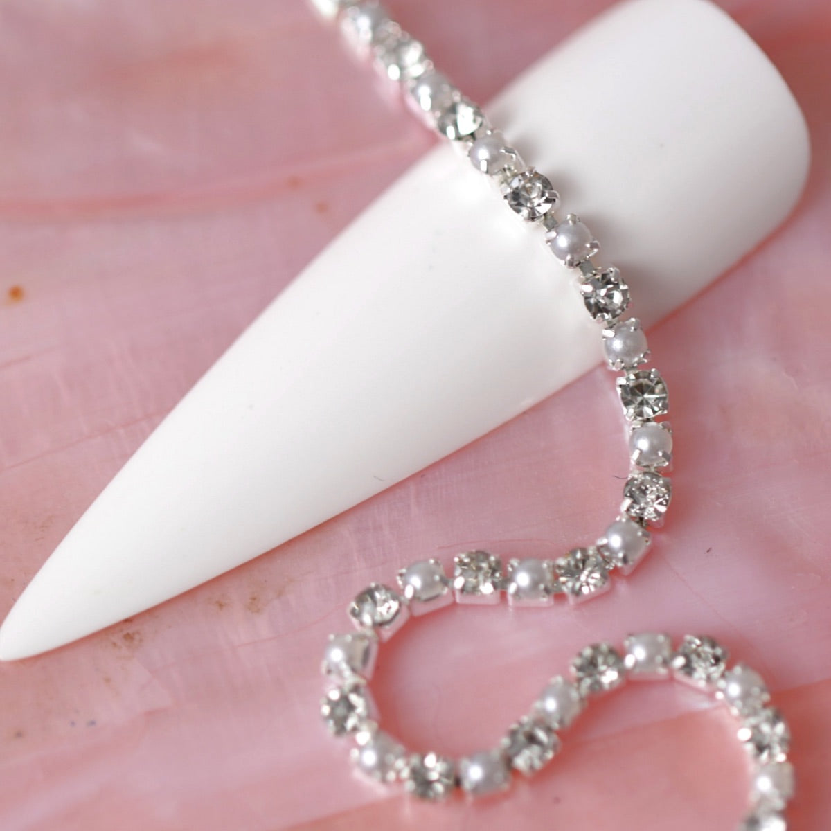 Silver rhinestone chain with alternating clear crystals and white pearls draped over a white nail tip on a pink background.