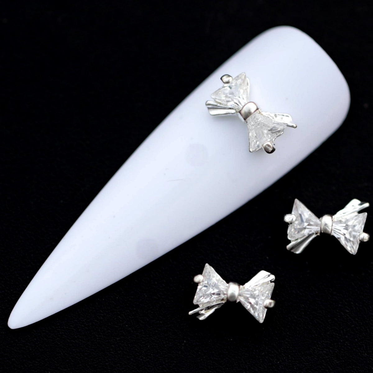 Silver bow‑shaped nail charms with crystal‑colored CZ stones on a white nail tip against a black background.