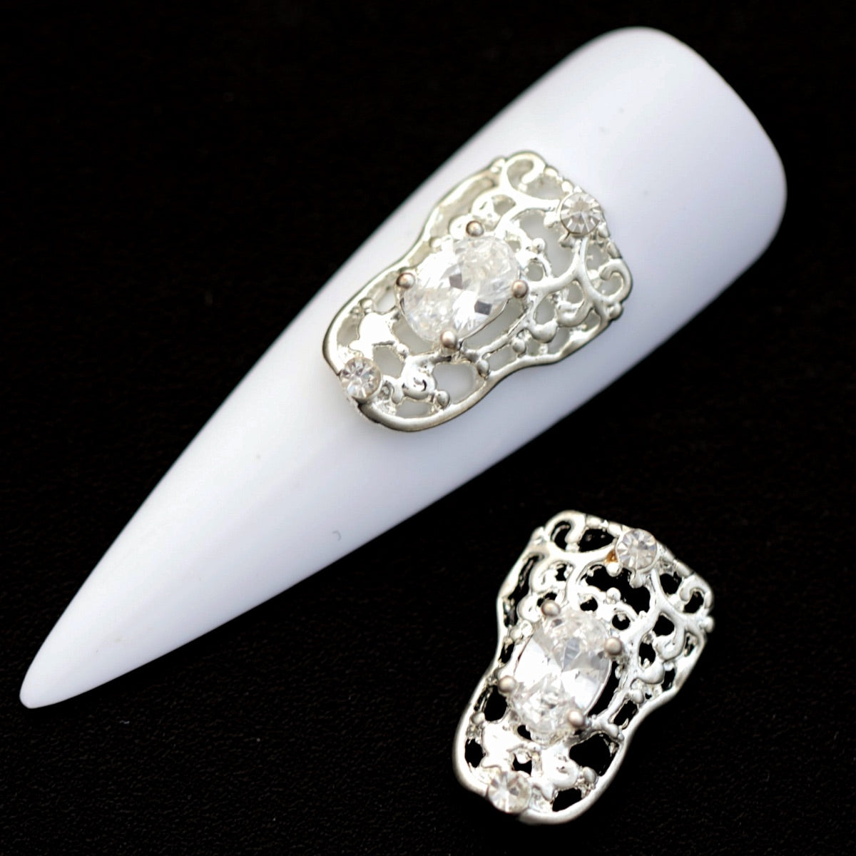 Silver filigree frame nail charms with central oval crystal CZ and small crystal accents on a white nail tip against a black background