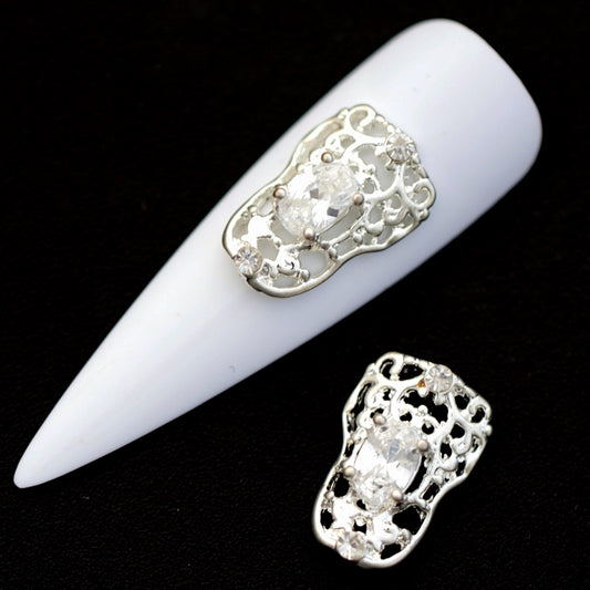 Silver filigree frame nail charms with central oval crystal CZ and small crystal accents on a white nail tip against a black background
