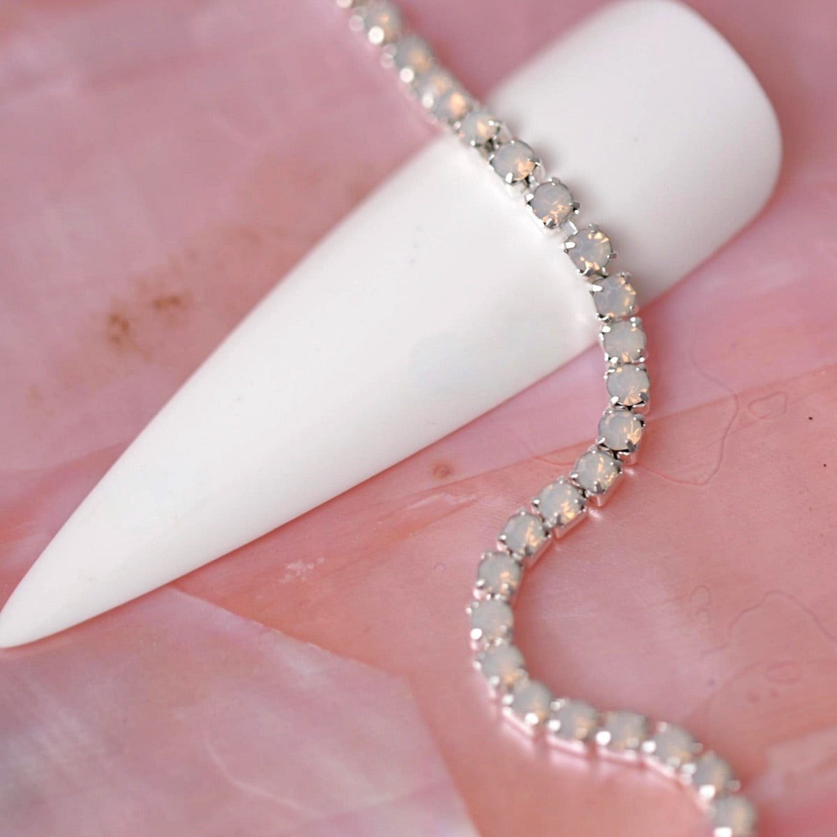 Silver rhinestone chain set with soft Light Rose Opal stones draped over a white nail tip on a pink background.