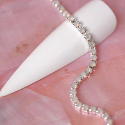 Silver rhinestone chain set with soft Light Rose Opal stones draped over a white nail tip on a pink background.