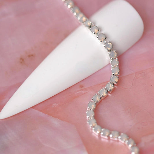 Silver rhinestone chain set with soft Light Rose Opal stones draped over a white nail tip on a pink background.