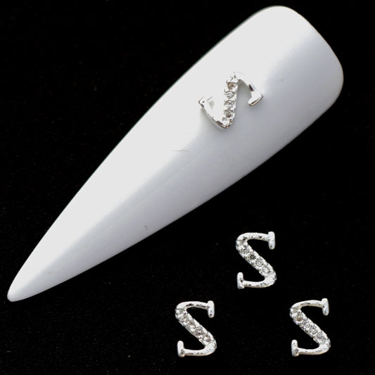 Silver S letter nail charms with crystal CZ accents on a white nail tip against a black background.