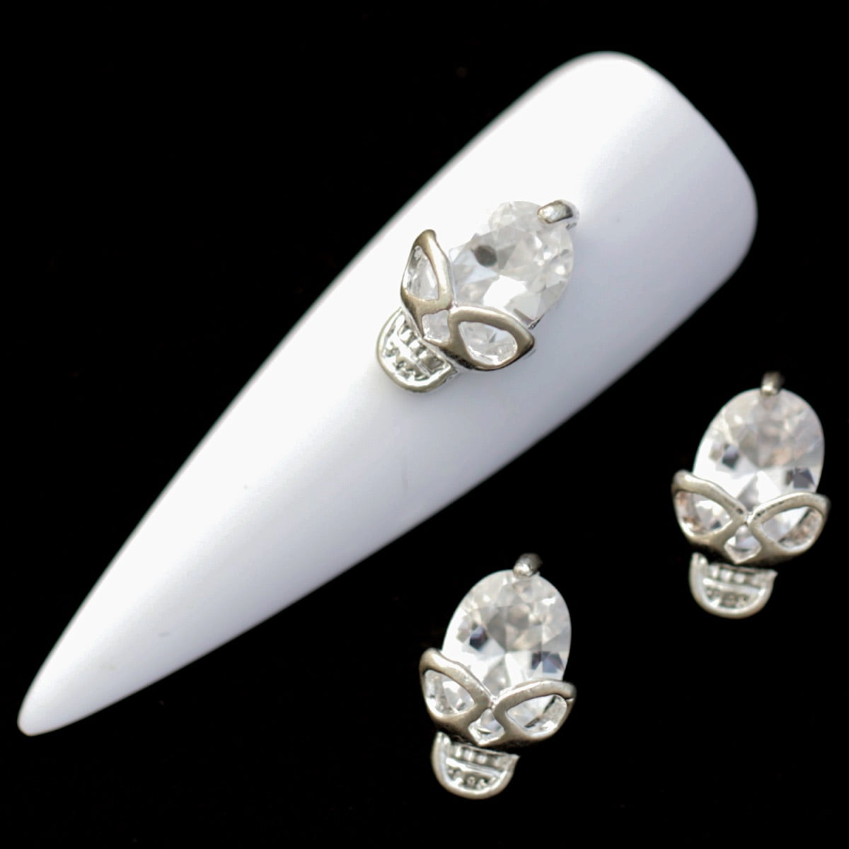 Silver skull nail charms with large crystal CZ skulls and metallic eye and teeth details on a white nail tip against a black background.