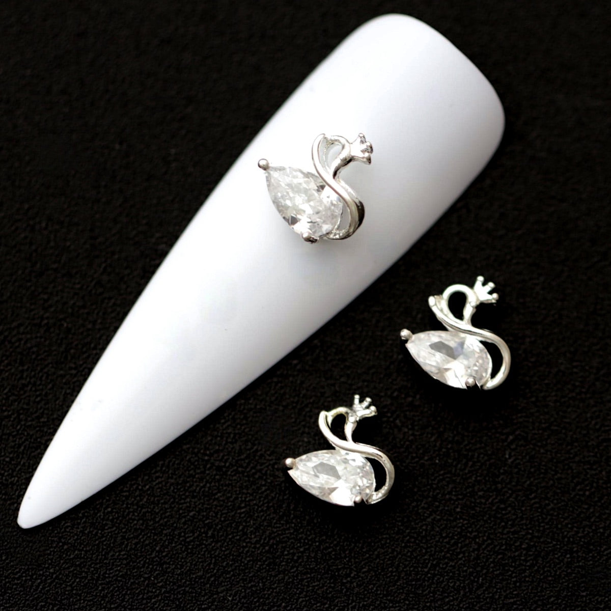 Silver swan nail charms with raindrop‑shaped crystal CZ bodies and tiny crown stones on a white nail tip against a black background