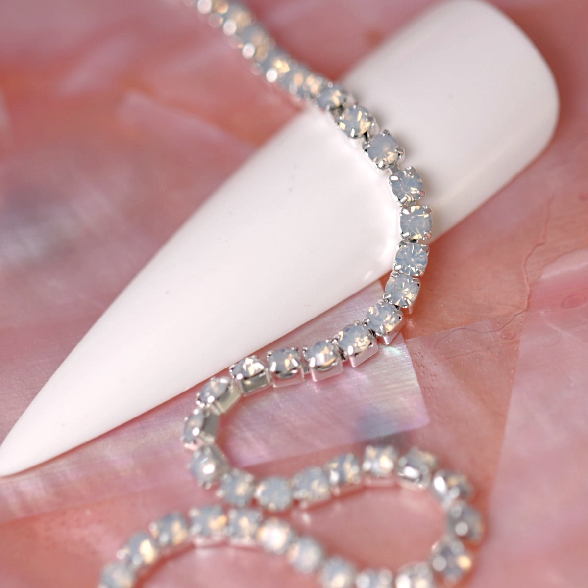 Silver rhinestone chain set with soft‑glowing White Opal stones draped over a white nail tip on a pink background.