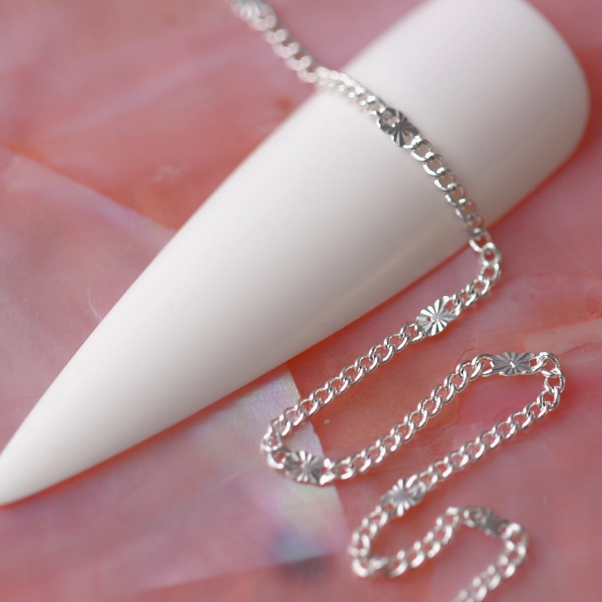 Ultra fine silver chain with tiny embossed sunburst discs draped over a white nail tip on a soft pink background.