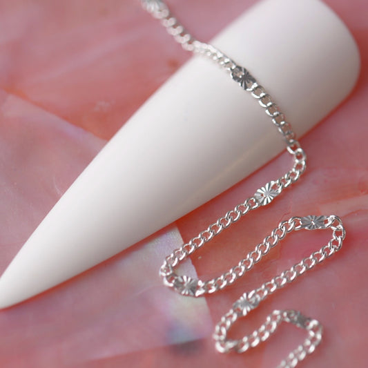 Ultra fine silver chain with tiny embossed sunburst discs draped over a white nail tip on a soft pink background.